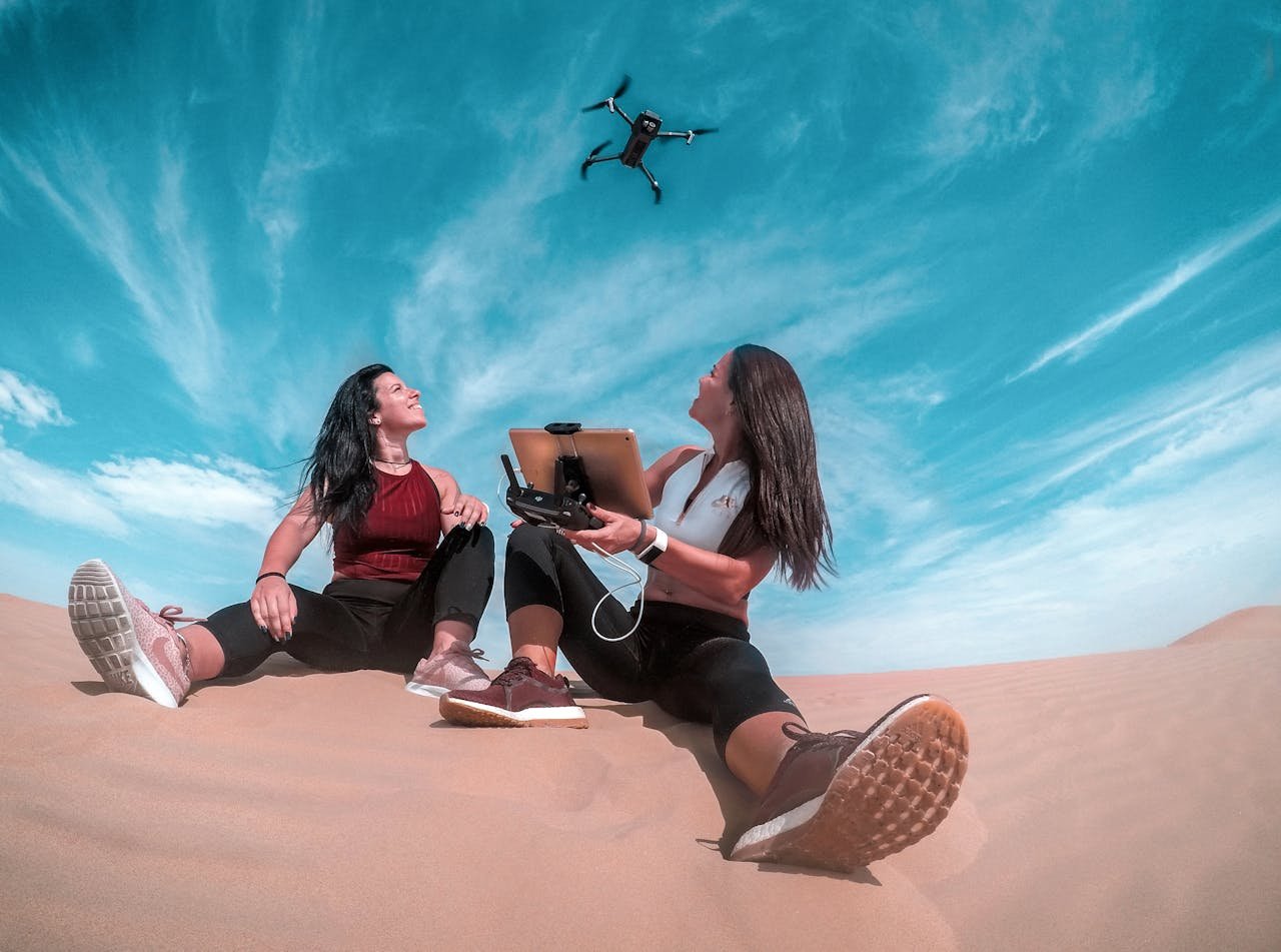 Two women on a Dubai sand dune control a drone under a bright blue sky.