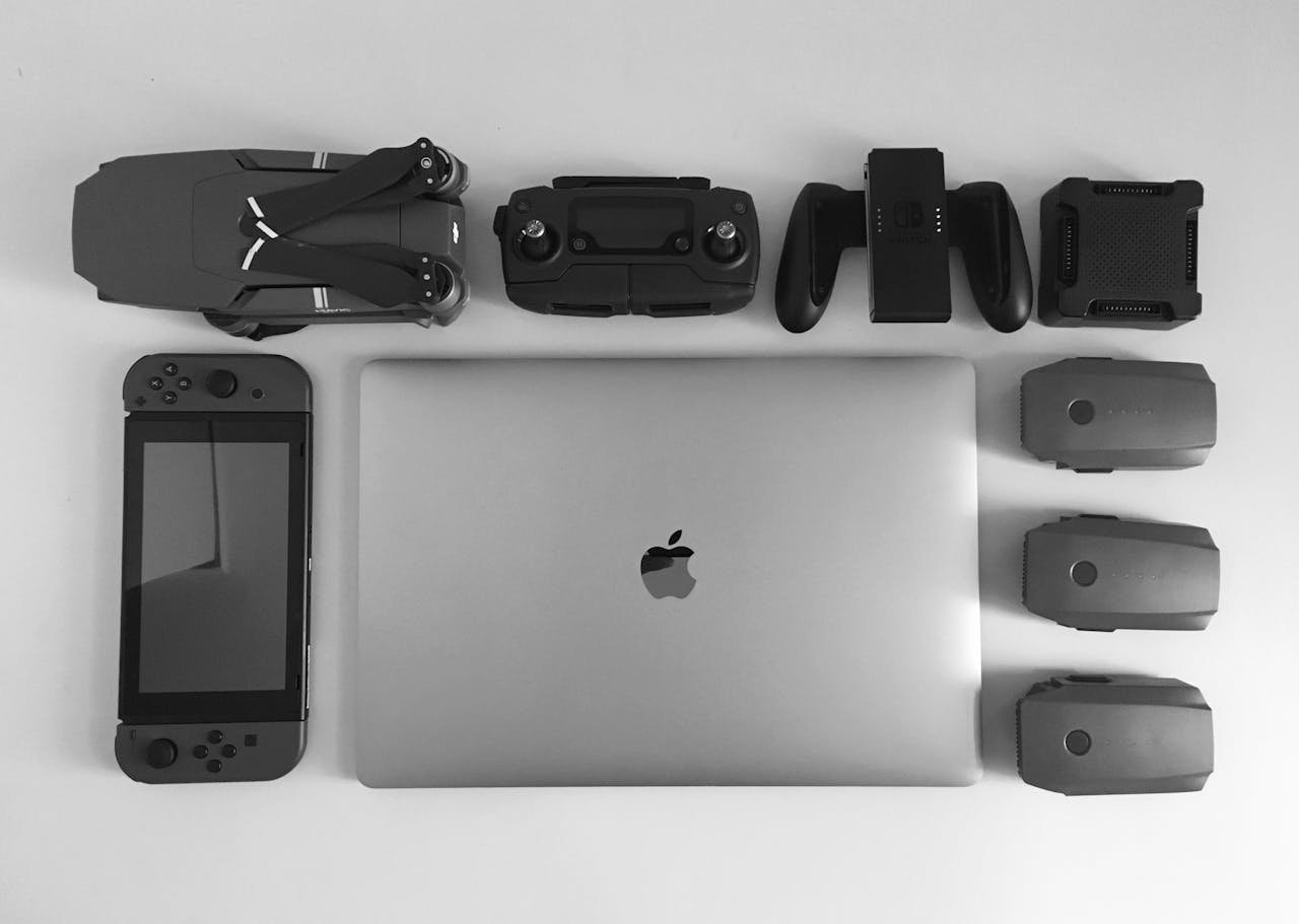 A black and white flat lay of various modern tech gadgets including a laptop, drone, and gaming device.